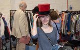 A woman wearing a red velvet top hat smiling at the camera. There are lots of rails of clothing behind her.