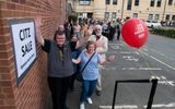 A long queue of people outside the stage entrance to the Citizens Theatre. A sign on the wall says 'Citz sale - queue starts here'. A red balloon is tied to the railing and people are smiling and waving at the camera.