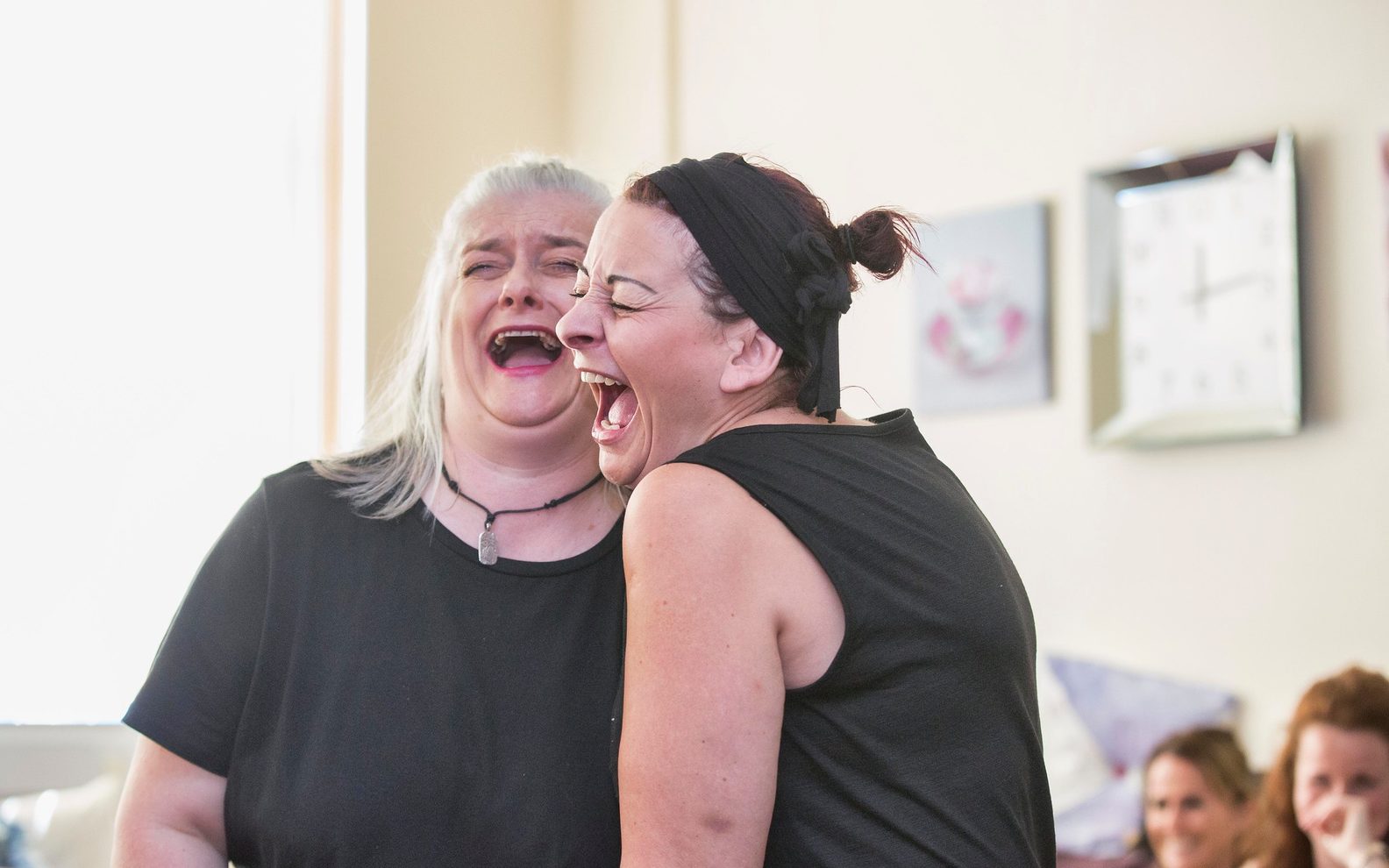 Two women laughing openly. They are both dressed in black and other women can be seen laughing in the background.
