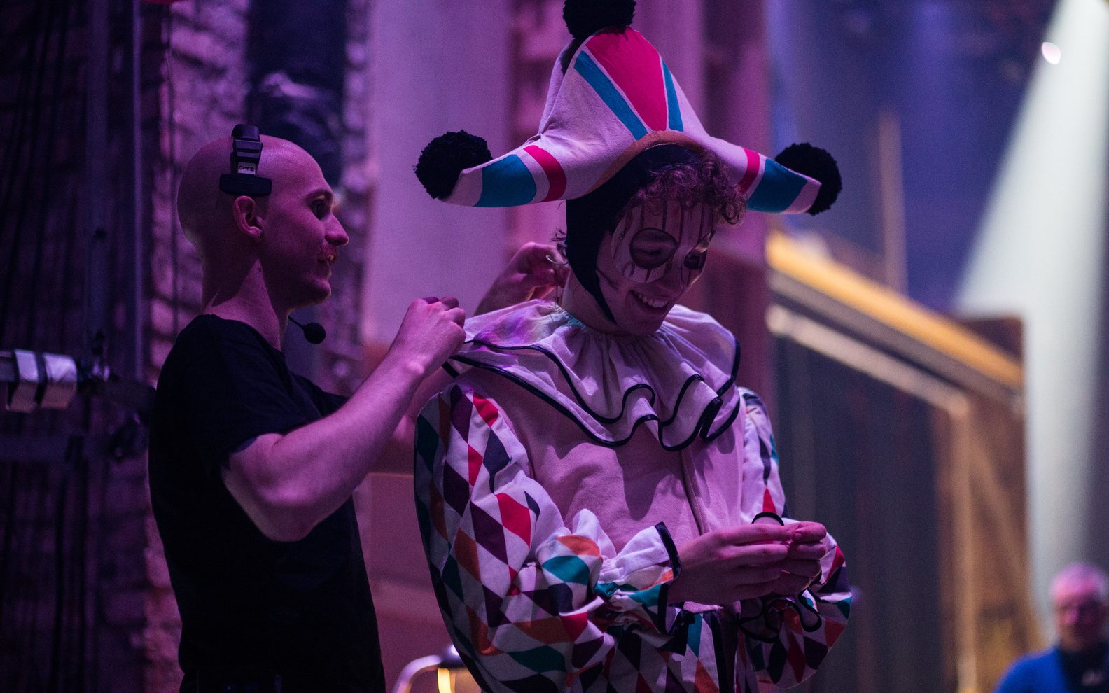 An actor stands backstage smiling in an Italian-style harlequin costume. A member of production staff is adjusting his microphone behind him.