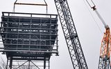 a large crane is pictured lifting a large metal structure into the air. The sky is blue with clouds.