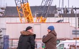 Two men in warm jackets and hats are standing next to each other. In front of them is a construction site with a large crane.