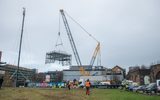 a large yellow and black crane is pictured lifting a large metal structure into the air. The sky is grey. In front of the crane are people in warm clothes, high visibility vests and hard hats