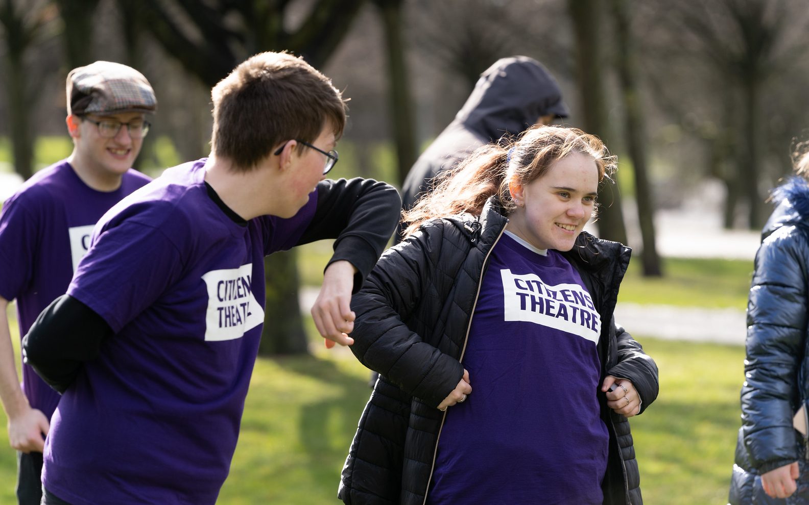 Young people from the Saturday Citizens group in a park. Two of them are in the foreground laughing, and all are wearing purple Citizens Theatre t-shirts.