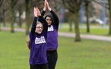 Two young people wearing purple Citizens Theatre t-shirts clap their palms above their heads. They are standing in a park.