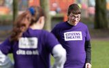 A young person with glasses and a purple Citizens Theatre t-shirt smiles while standing in a park. Another young person stands in front of them with their backs to the camera.