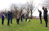 A group of young people dressed in winter clothes stands in a park facing two adults. All are raising their arms above their heads.