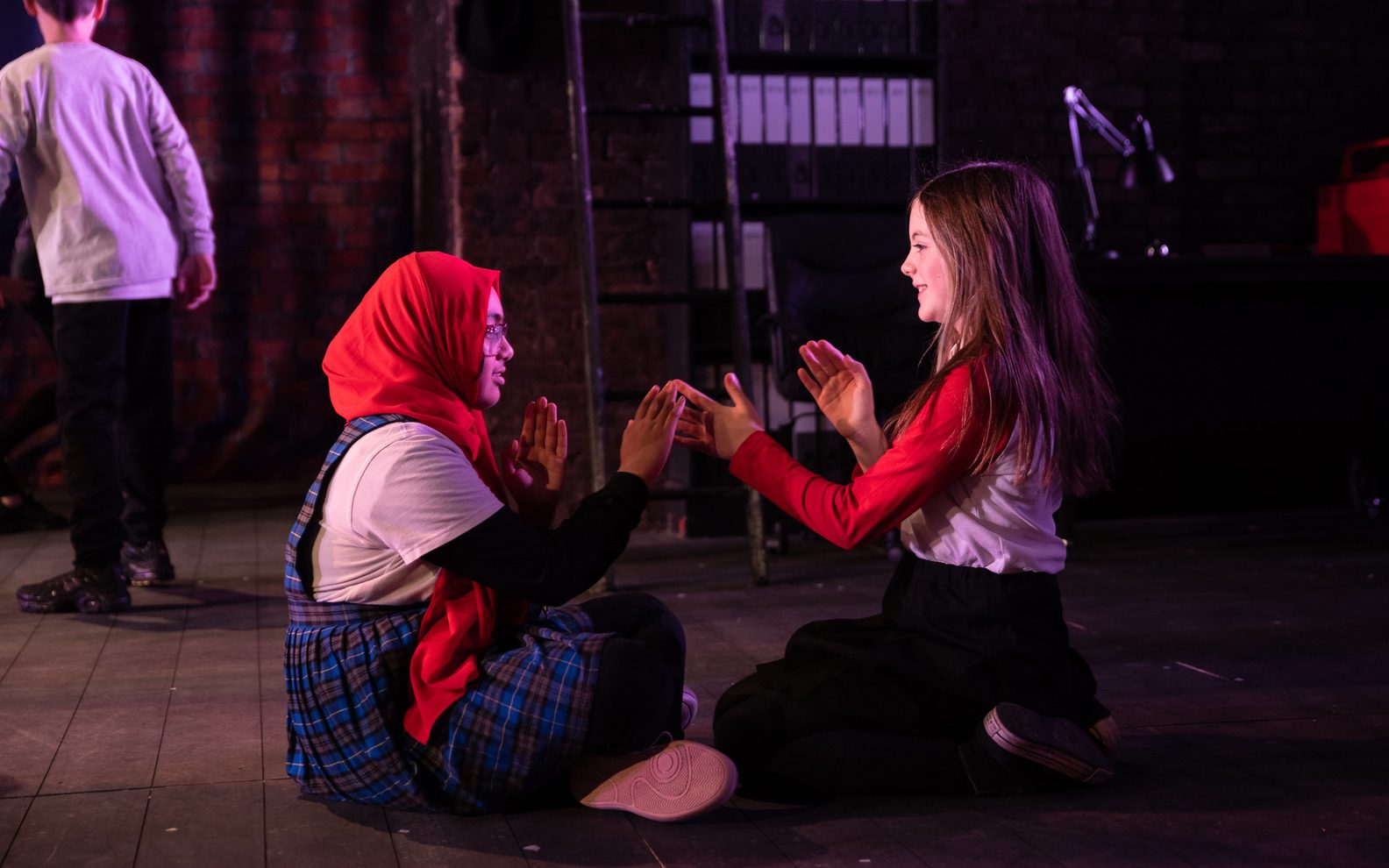 Two primary pupils sitting on a theatre stage. The girls are facing each other and playing a hand clapping game.