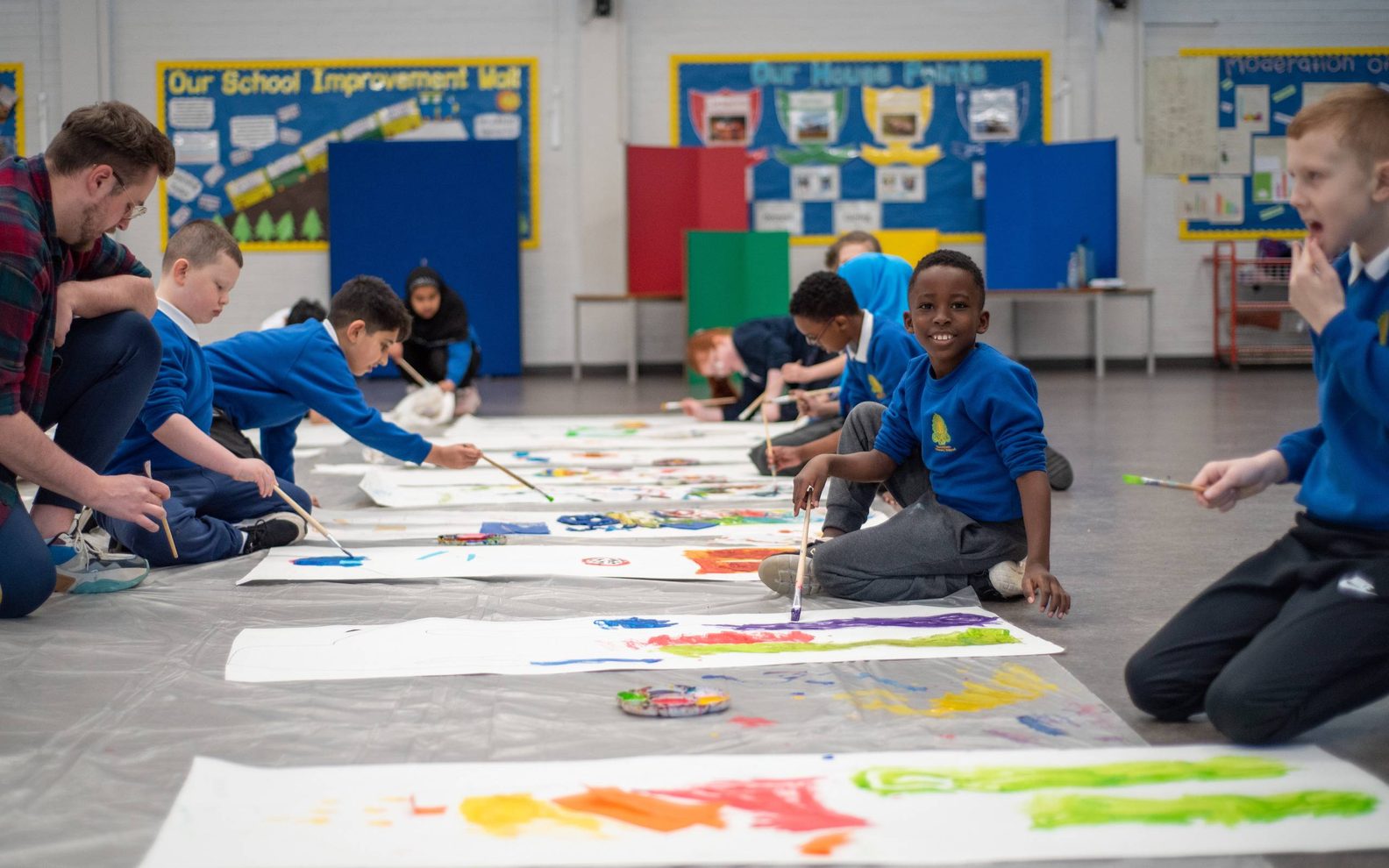 A group of primary pupils painting in bright colours on large sheets of paper on the floor of a school hall. They are wearing blue school uniforms.