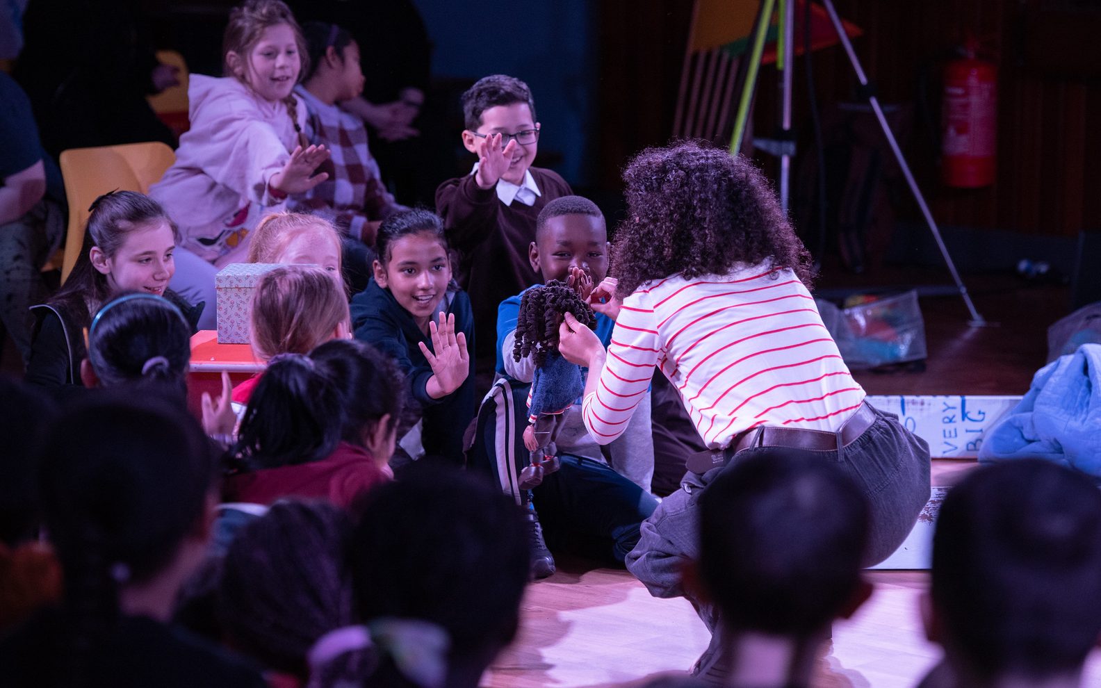 A performance of Maud’s Map at St Francis Primary School. An actor is holding a puppet and an audience of primary school pupils are sitting watching with their arms outstretched.