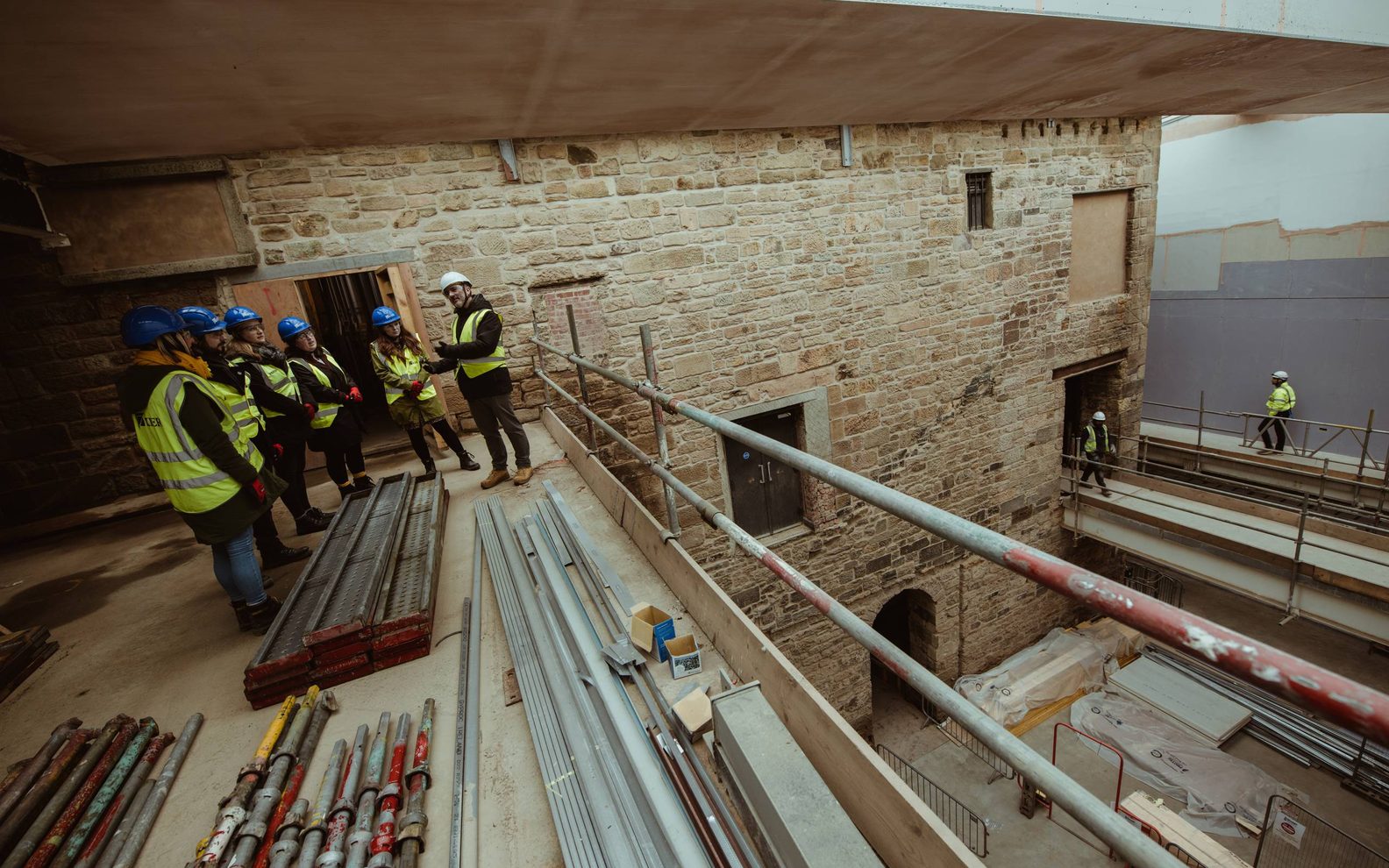 Six people on a tour of the Citizens Theatre building redevelopment. They are dressed in high vis vests and hard hats. They are standing on a balcony with scaffolding around them.