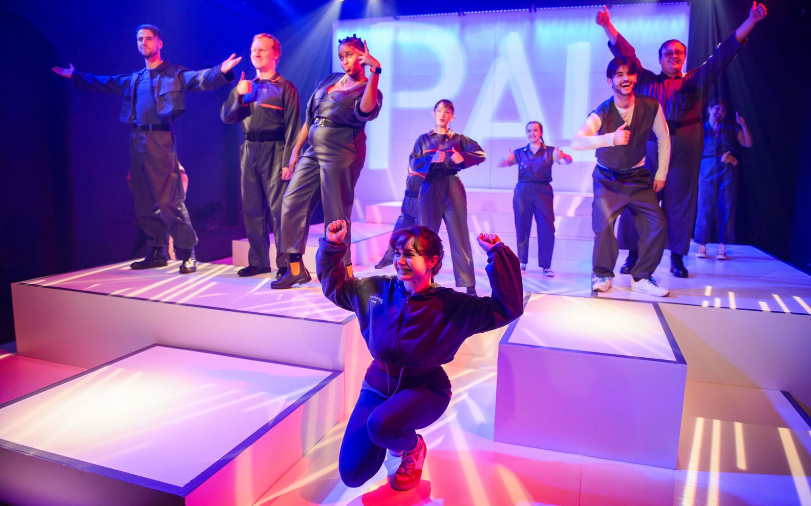 A group of 9 young people are standing on a futuristic stage. They are all wearing grey boiler suits. They are standing in a variety of poses. Some are standing with their thumbs up, other have their arms stretched up, others are crouched down and smiling.