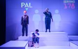 A stage show. A woman is sitting on steps wearing a sling on her arm. A woman is stood behind her holding a bandage to her head. There is a man to the side of them. The word PAL is projected on the wall behind them.