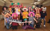 A group of learning disabled adults standing together and smiling and posing for the camera. They are all wearing colourful hats and oversized theatrical props.