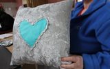 A person holds up a grey pillowcase with a turquoise heart stitched onto it.