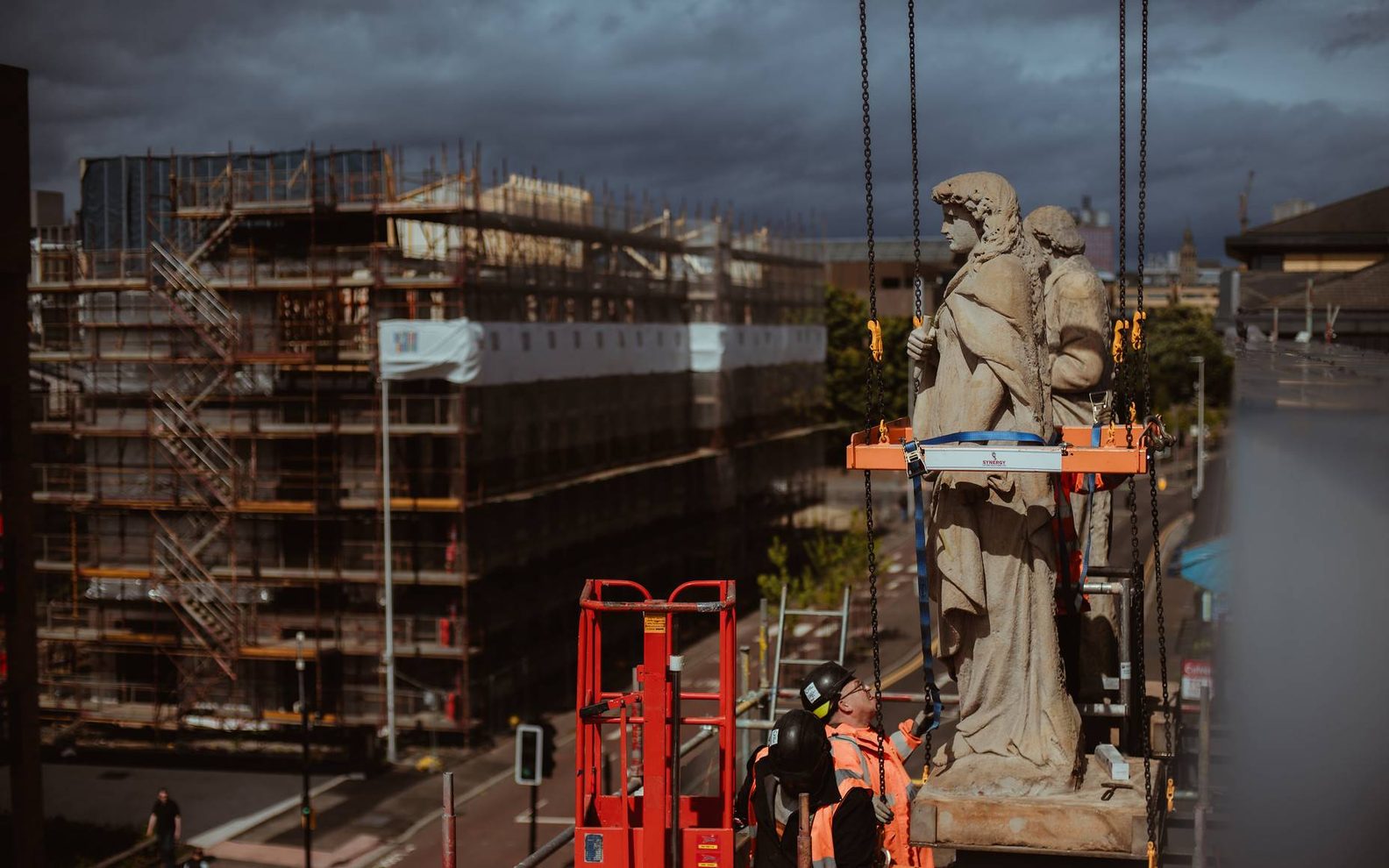 The Citizens Theatre redevelopment building site, two men in high-vis clothing are standing on scaffolding in front of three historic statues on the roof of the theatre. In the background, you can see a main road and construction across the road.