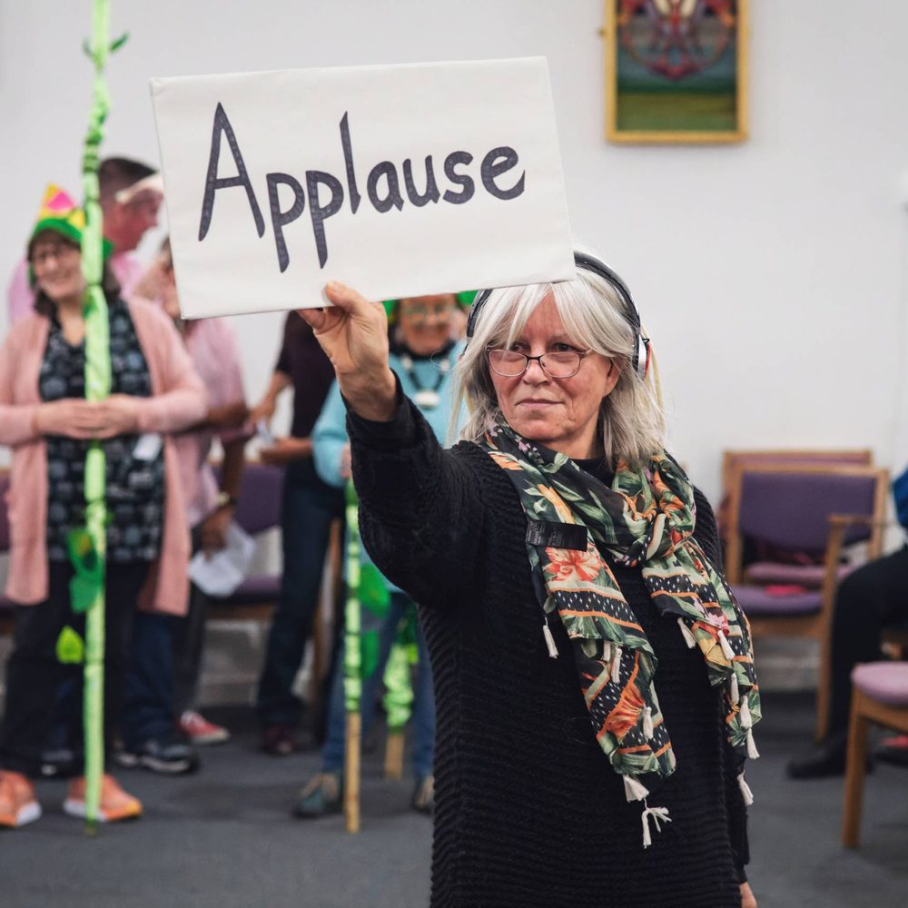 A woman holding out a sign that has the word ‘Applause’ written on it. A group of people are standing in the distance behind her wearing theatrical accessories and holding props in their hands. To her right people are sat in chairs holding scripts in their hands.