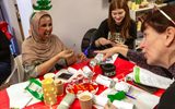 Three women sit at a table doing Christmas-themed crafts. One is wearing a headscarf and a Christmas tree headband and laughing.