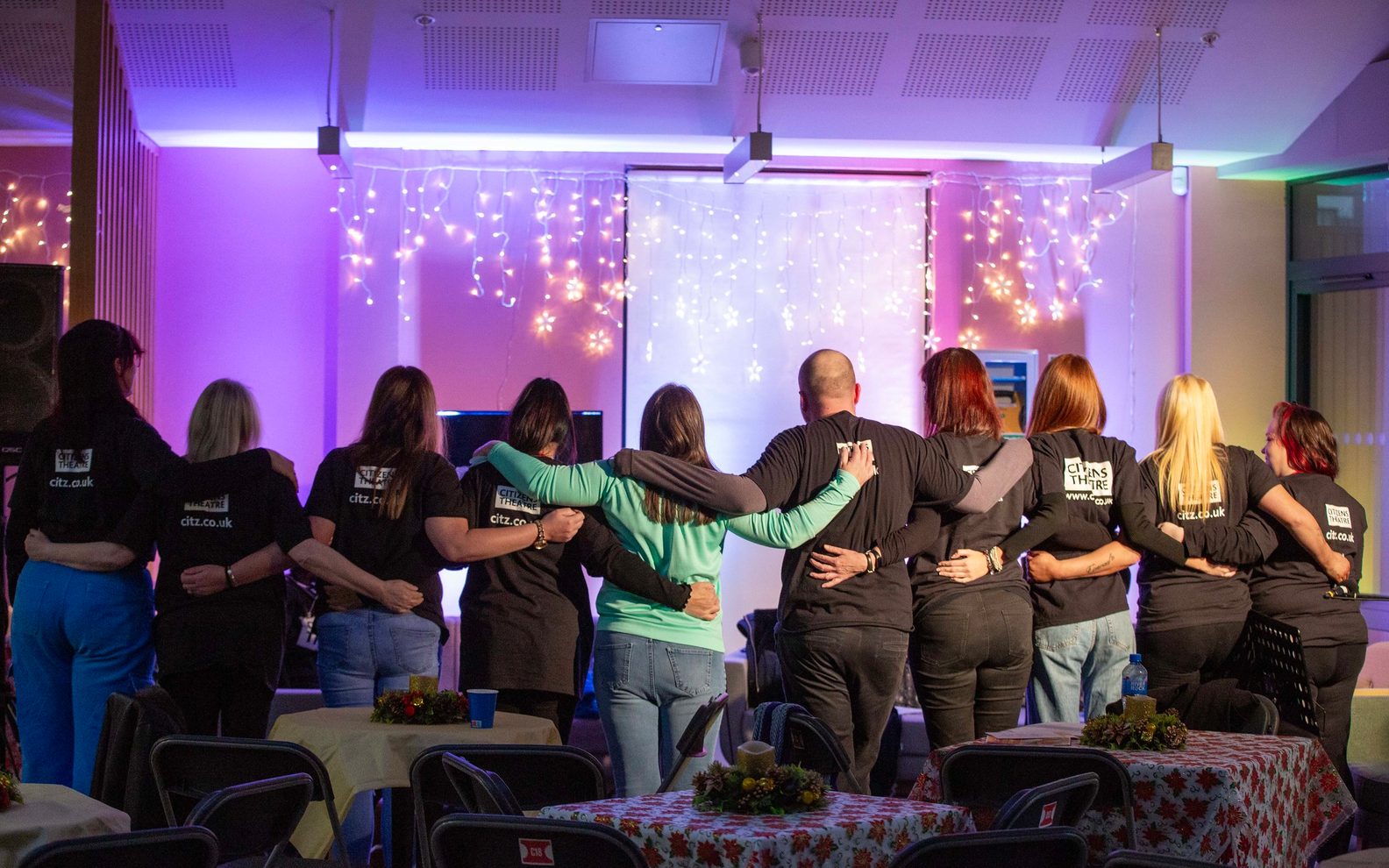 A group of people standing in a line, seen from the back. They are all holding each other and wearing Citizens Theatre t-shirts. The room is illuminated in purple lighting with fairy lights.