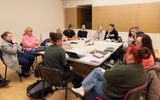A large group of people are sitting around a table. They are listening to somebody presenting information from a laptop.