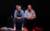 A performance image. Two young women sit on stage and talk to each other.