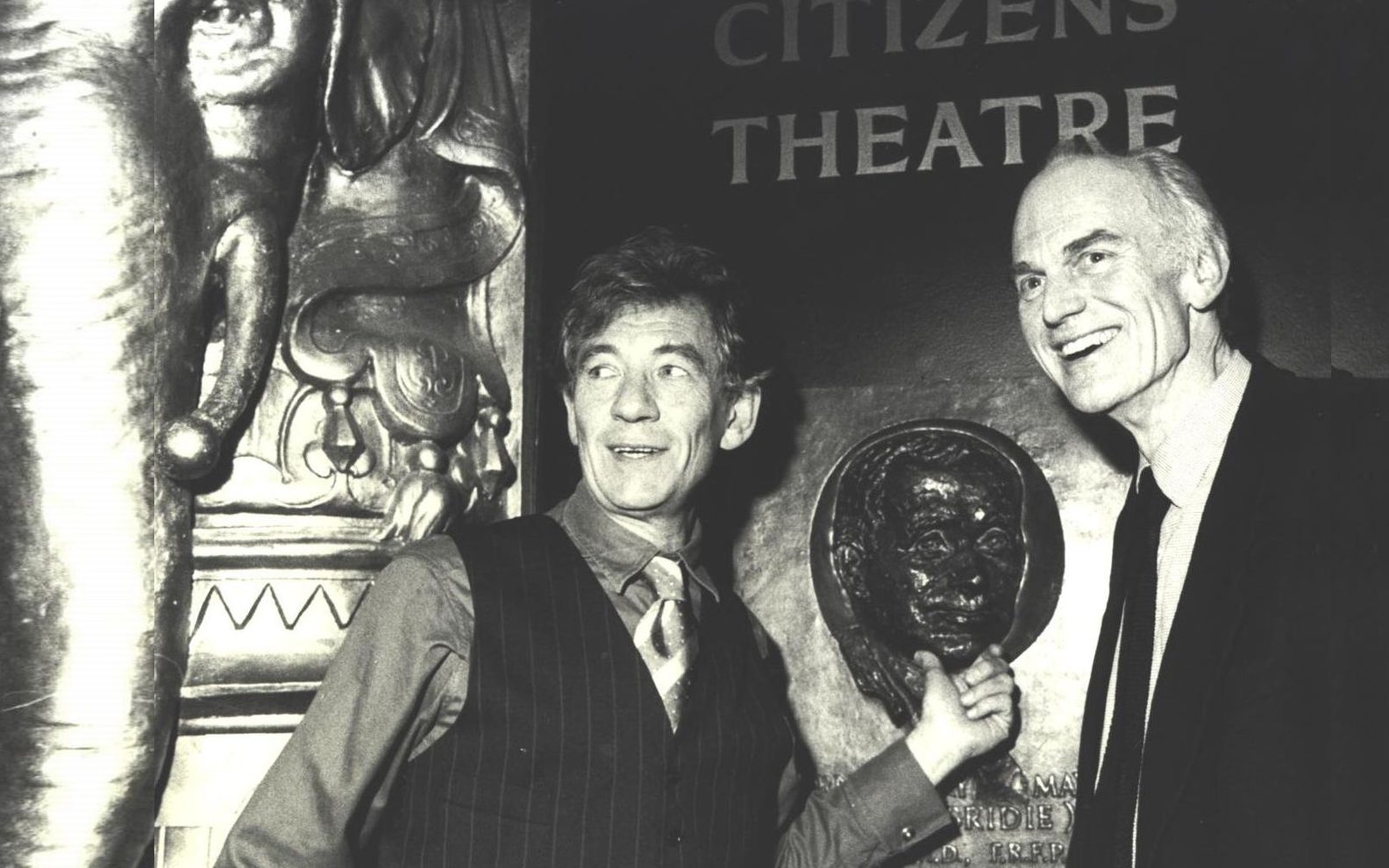 Sir Ian McKellen and Giles Havergal pose in the Citizens Theatre lobby. The name of the theatre and a plaster elephant head are seen behind them.