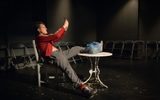 A production shot. A man in a tracksuit lounges on a chair and looks at a phone in his hands.