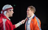 A production shot. Two men in bright tracksuits face each other. One holds a wooden gavel under the other's chin.