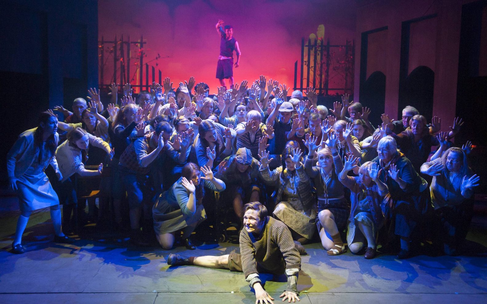 A large number of actors on a stage are facing the camera, crouching down and raising their hands to the sky. They are dressed in a variety of drab clothes from the 1950s. One actor is lying on the ground in the centre of the stage with the other actors gathered round him. He has a grimaced face. Another actor is standing at the very back of the stage. He is standing on a wall above the others looking out to the camera with one hand in the air.