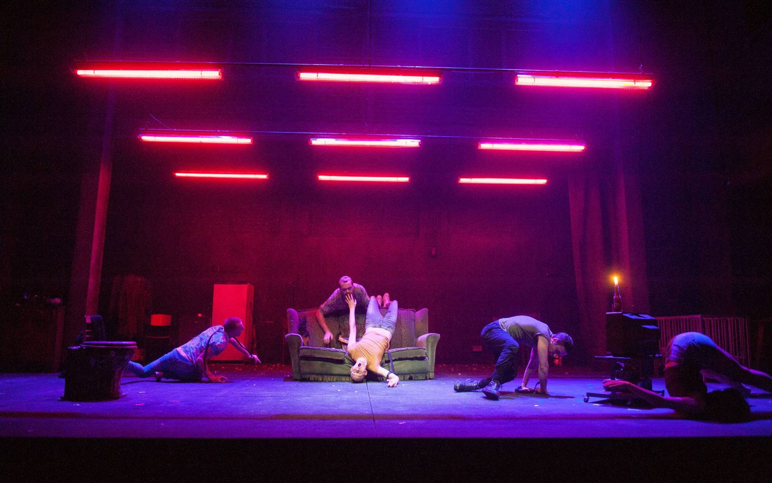 There are three people on stage who are all crouched or rolling on the floor. Two more people are draped over a sofa. Red neon lights hang from the ceiling.