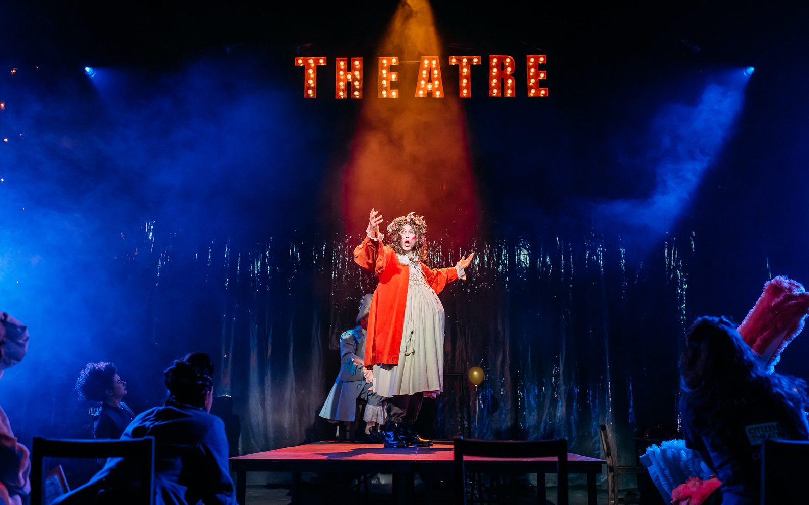 A theatrical production. A man stands on a platform with a spotlight on him. He is talking animatedly. He has lots of make up on and is wearing a white gown and red robe.