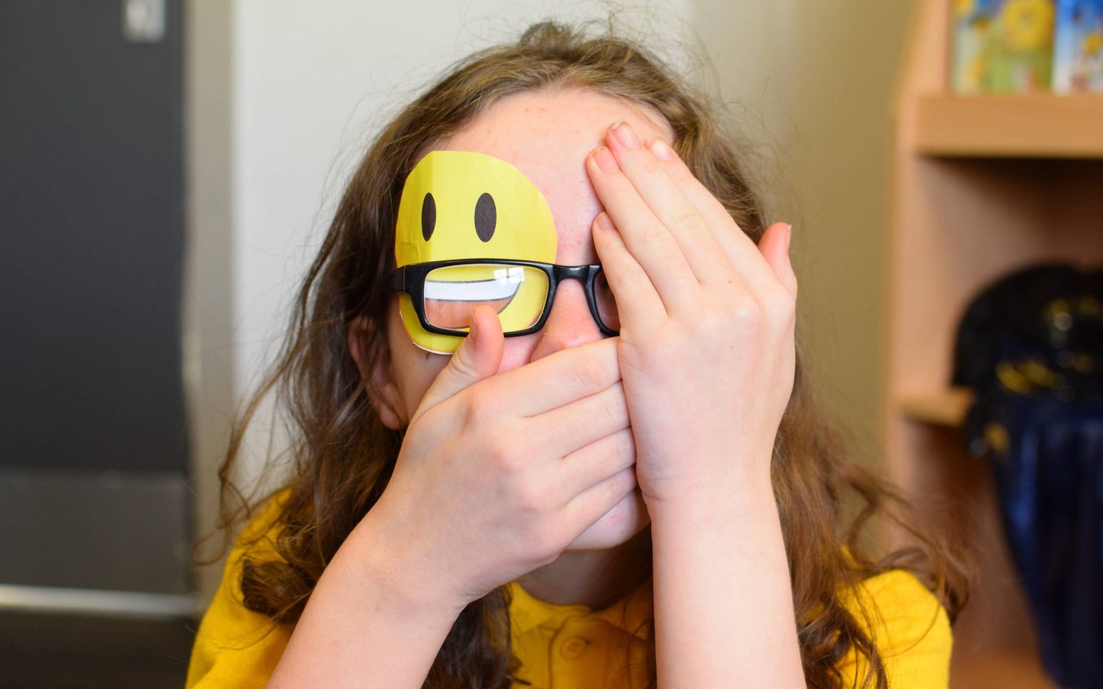 A young girl is looking at the camera. She has a yellow smiley face covering one eye. She is covering the other eye with her hand. She is covering her mouth with her hand.