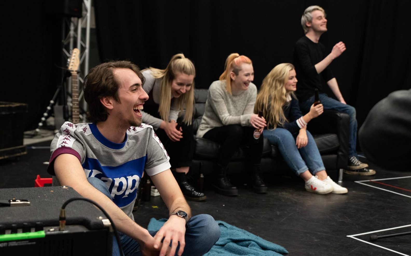 Five young actors, two male and one female. They are sitting on a sofa on the set of a stage. They are looking off to the side and laughing.