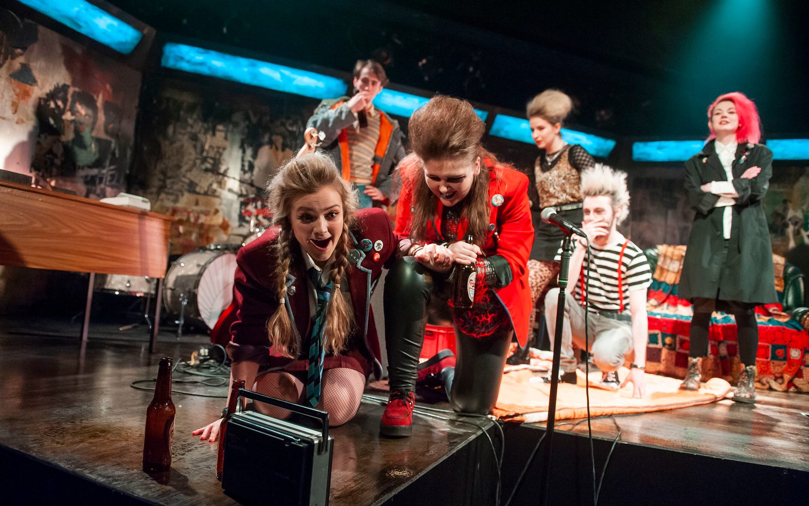 WAC Jaggy Nettles A highly theatrical production image of a show. Everybody is wearing punk style clothing. They are on a stage with graffiti style surroundings. A group of young people are crouched round an 70s style radio.
