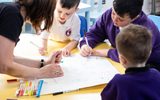 Three young schoolboys are standing round a yellow school desk, they are wearing white school t-shirts and purple school jumpers. They are next to a member of Citizens Theatre staff with short brown wavy hair who is wearing a black t-shirt. They are leaning over a large white sheet of paper and using colourful felt tip pens to write and draw.