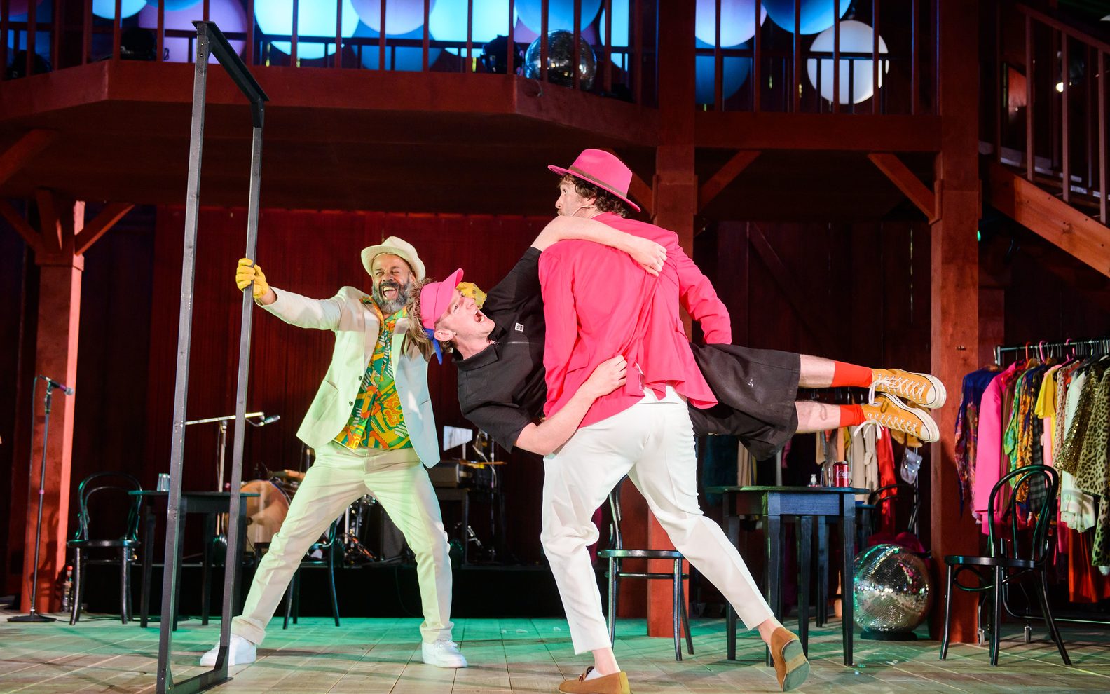 A theatrical production. Three men in bright clothes are on stage. One is holding a door frame while another is holding the third man horizontally and walking towards the frame.