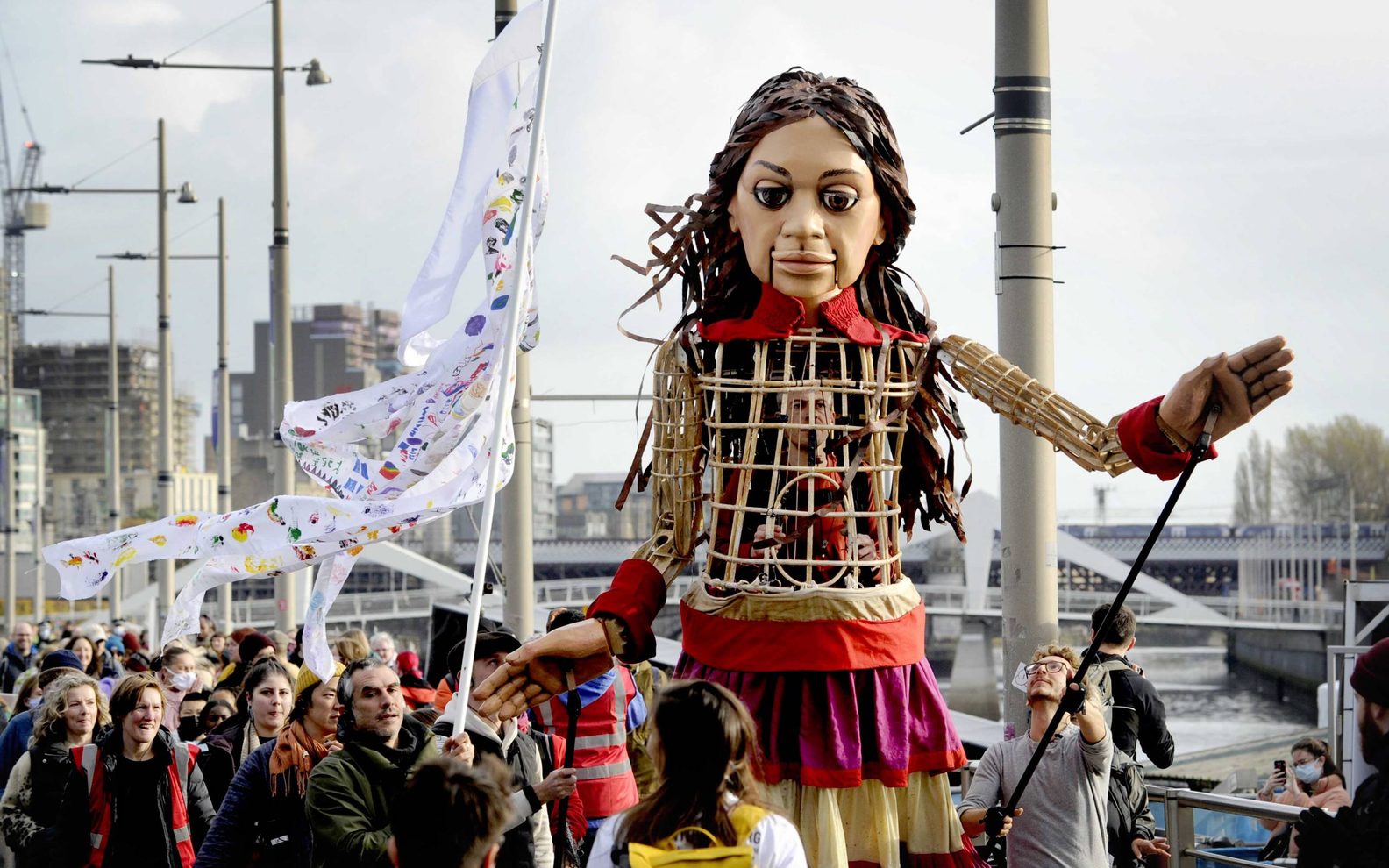 A large wicker puppet of a young girl walks along the street in Glasgow. She is surrounded by people who are looking at her and waving flags.