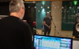 A man stands in a recording booth with headphones over his years. Another man can be seen out of focus in the foreground, mixing the track.