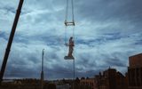 A statue hanging on a crane as it is moved. It is in stark contrast with the sky behind it.