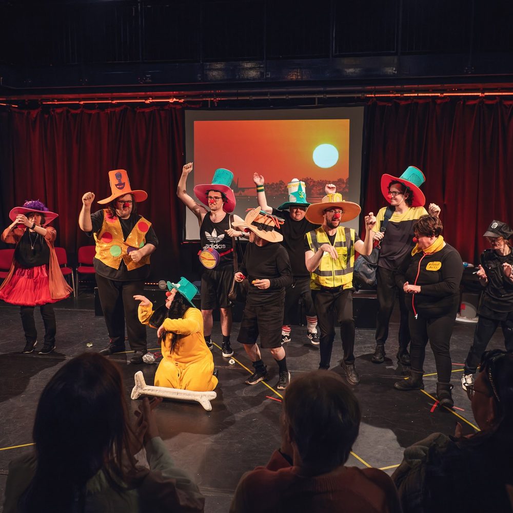 A large group of people standing on a stage. They are all wearing large hats, red clown noses and holding colourful props. In the foreground, you can see the backs of an audience .