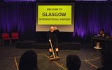 A stage show. A man dressed in black and a red clown nose is sweeping the floor. A sign behind him says 'Welcome to Glasgow International Airport'. In the the background a woman is sitting at a desk reading.