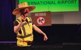 A stage performance. A man is wearing a large hat, red clown nose and high vis vest. He is smiling. A screen behind him has the words 'International Airport' on it and there is a large 'No dogs' sign in front of it.