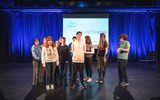 A group of young people standing together on a stage. Words are projected on a screen behind them including 'I love fashion' and 'I value my friendships'.