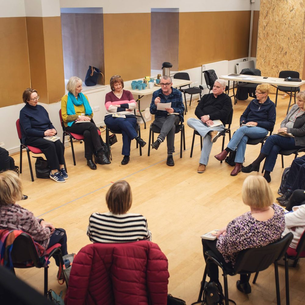 A play reading session. A group of older adults sit in a circle, holding books.