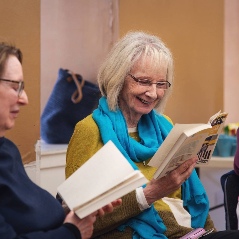 A play reading session. A woman with glasses and a bright blue scarf smiles while reading from a book.
