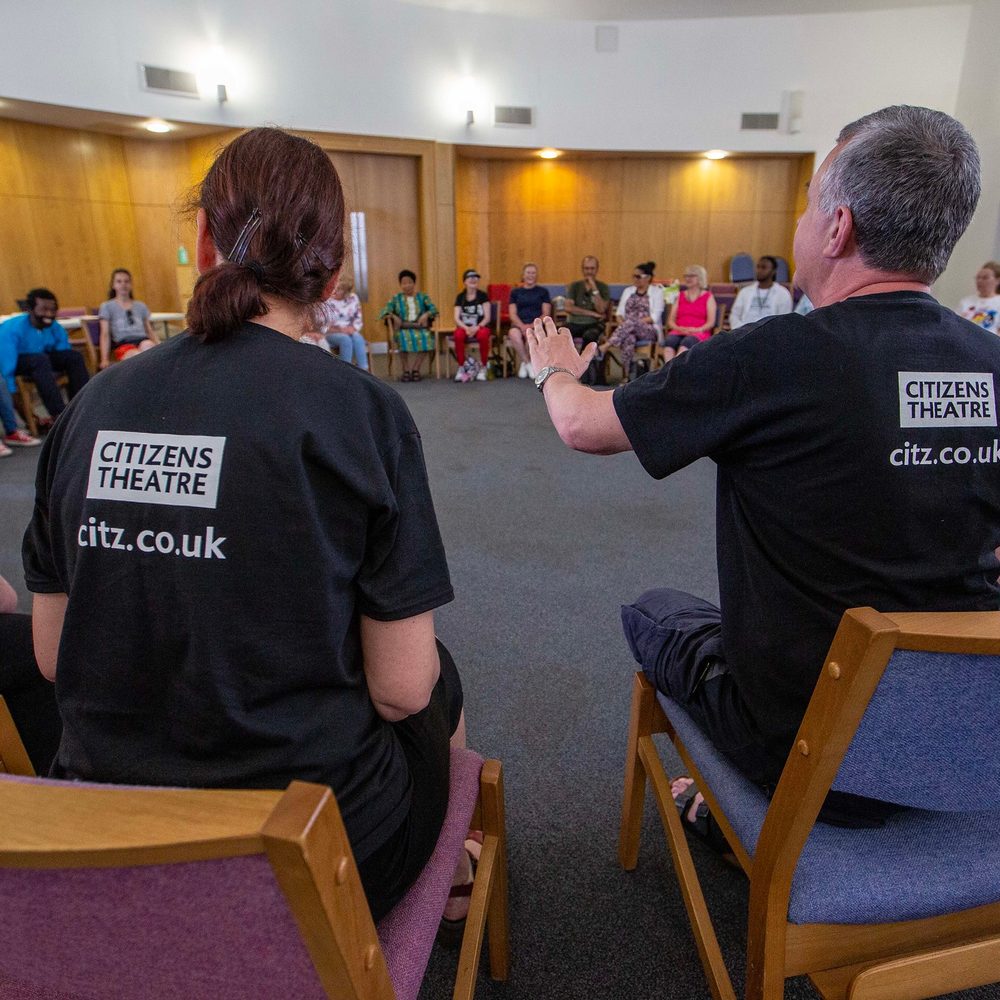 Com Co at Refugee Festival Scotland 2023 A large group of people sitting in a circle. The two people in the centre of the image are wearing Citizens Theatre t-shirts.