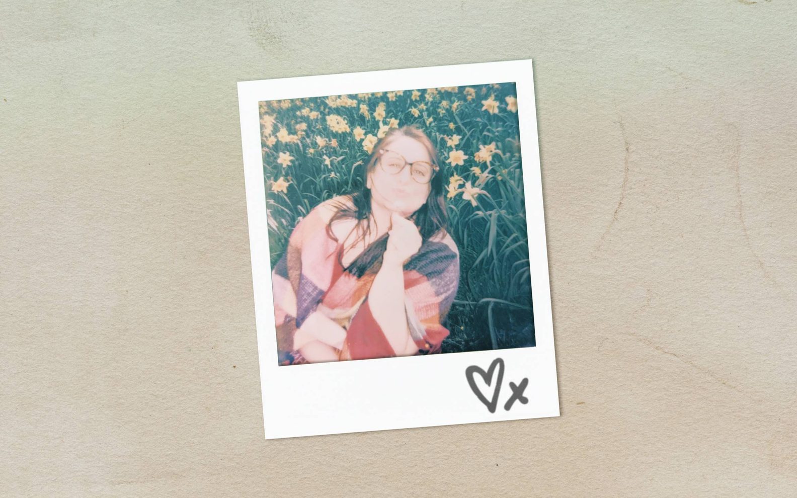 A faded polaroid picture of a young woman in glasses sitting in a field of flowers. A heart and 