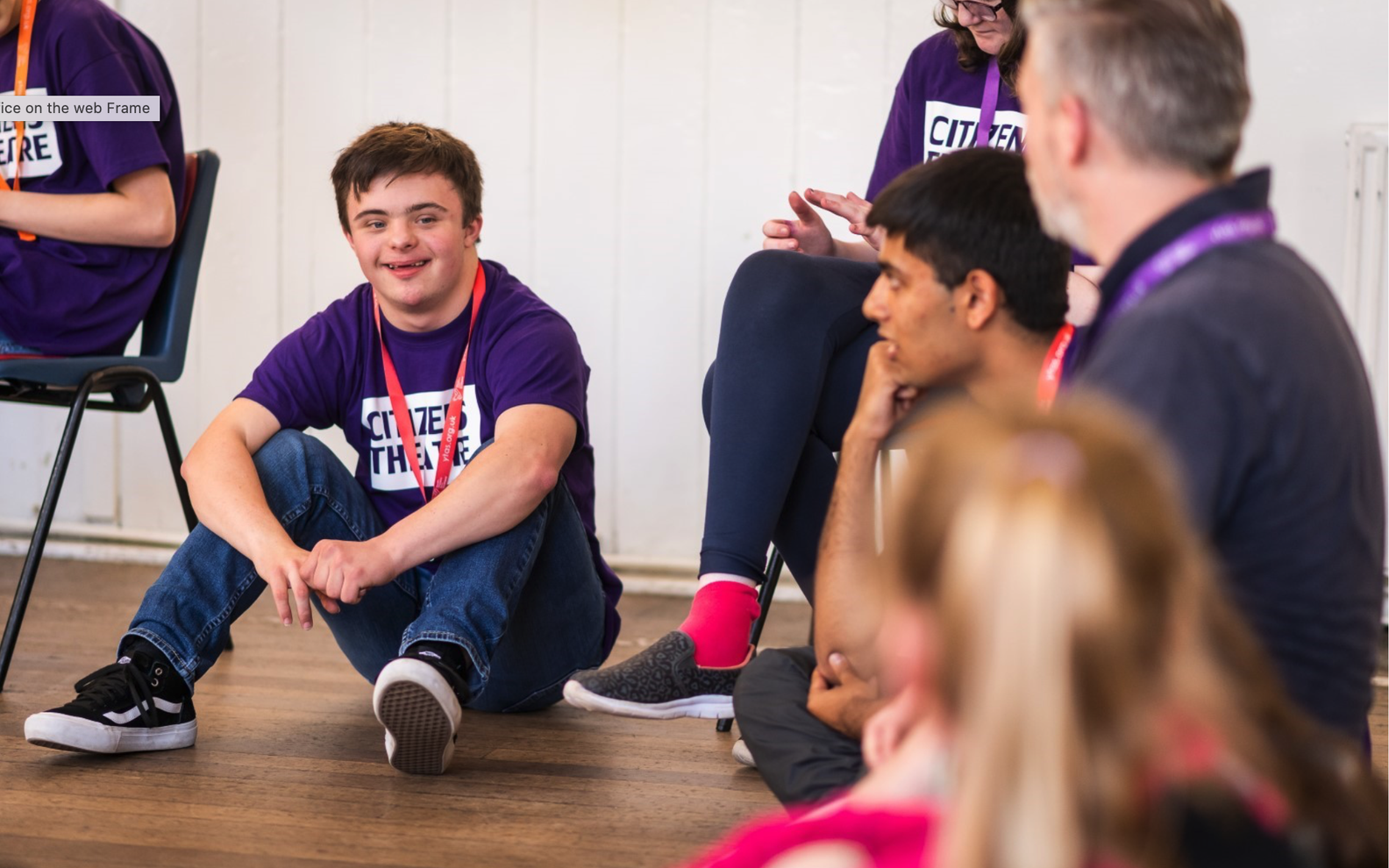 A group of learning disabled young people sit on chairs and on the floor. In focus, a young man in a purple Saturday Citizens listens to an instructor speak.