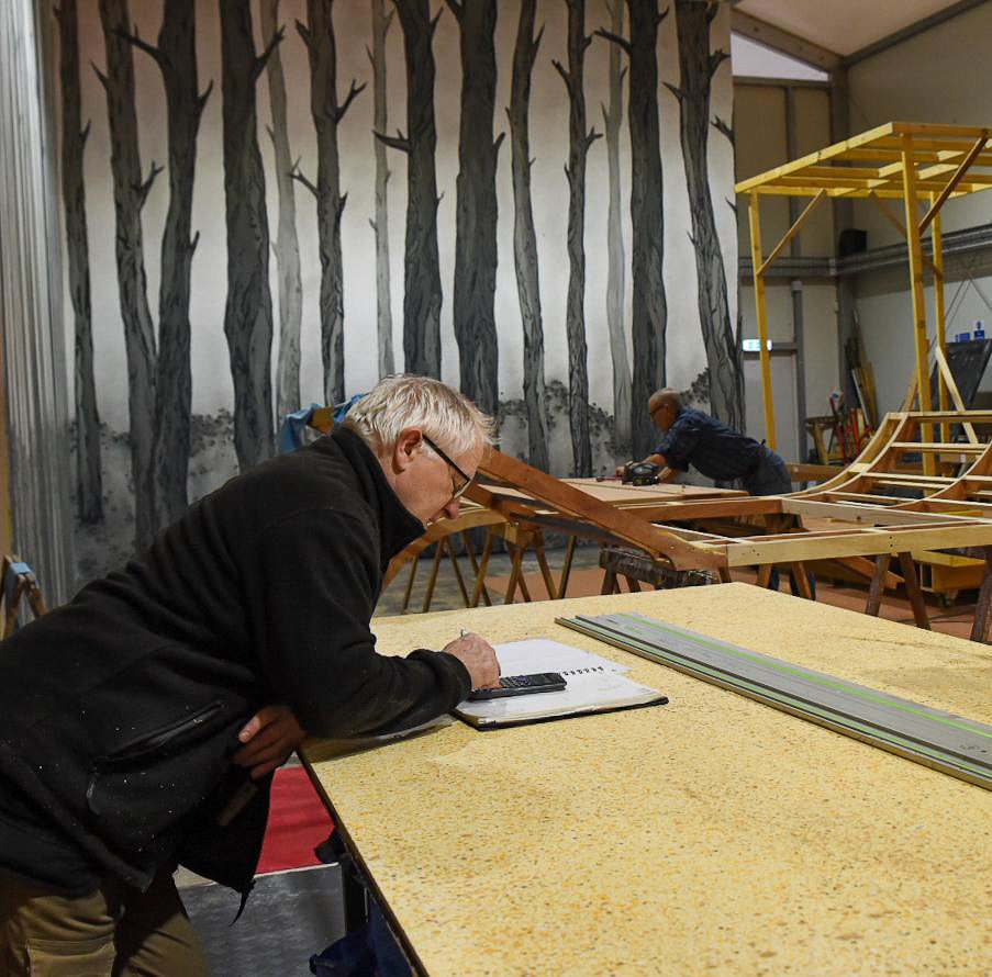 Denis Murphy in his workshop. A large painting of trees is visible in the background. He is looking at a calculator and notepad.