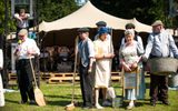 A group of performers at an outdoor event. They are wearing old fashioned clothing and holding cleaning products like brooms, mops and wash baskets.